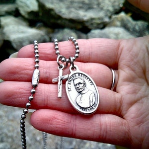 May include: A silver chain necklace with two charms. One charm is a cross and the other is a round medal with a picture of a man and the text "St. Andre Bessette Pray For Us".