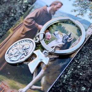 May include: A silver keychain featuring a St. Francis medal and a cross charm. The keychain is attached to a card with an image of St. Francis and a wolf. The words "SAINT FRANCIS" are printed on the card.
