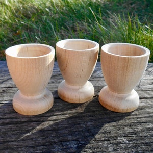 May include: Three wooden egg cups, each with a rounded top and a flared base, are arranged in a row on a weathered wooden surface. The cups are made of light-colored wood and have a natural finish.