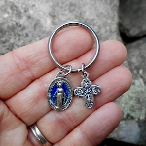 May include: Silver keychain with a blue enamel oval charm featuring the Virgin Mary and the words "Miraculous Original Consecration" and a silver cross charm with floral details.