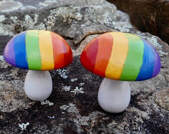 May include: Two rainbow painted mushroom figurines on a gray rock surface. The mushrooms have white stems and are painted in the colors of the rainbow.