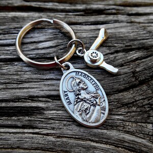 May include: Silver key chain with a silver charm of Mary Magdalene. The charm has the words "Mary Magdalene" inscribed on it. The key chain also has a silver charm shaped like a hair dryer.