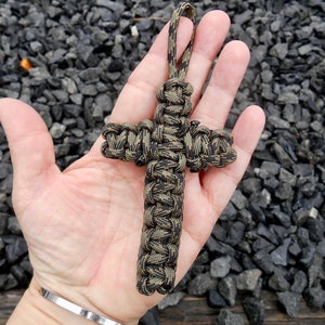 Puede incluir: Un llavero de cruz de paracord de camuflaje negro y marrón. La cruz está atada con un patrón de nudos.