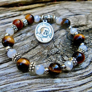 Puede incluir: Un brazalete de cuentas marrones y blancas con un dije de plata con una imagen de San Pío. Las cuentas están hechas de ojo de tigre y ágata blanca.