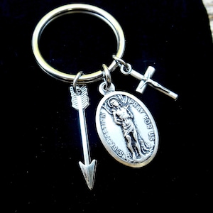 May include: Silver keychain with a Saint Sebastian medal, an arrow charm, and a cross charm. The medal features an image of Saint Sebastian with the text "Pray for us" and "St. Sebastian" around the edge.