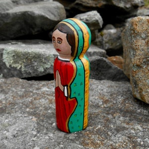May include: A hand-painted wooden doll figurine of a woman wearing a red dress and a green and gold patterned shawl. The doll is standing with her hands clasped in prayer.