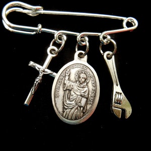 May include: Silver safety pin with three charms: a cross, a medal of Saint Vincent Ferrer, and a wrench. The medal has the text "St. Vincent Ferrer Pray For Us".