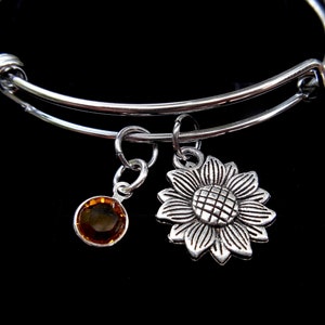 May include: Silver-toned adjustable bangle bracelet with two charms. One charm is a sunflower, the other is a round, amber-colored gemstone. The bracelet is displayed against a black background.