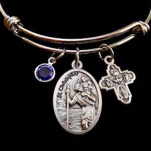Könnte beinhalten: Ein silbernes Armband mit drei Anhängern: einem blauen Kristall, einem silbernen Kreuz mit religiösen Motiven und einem silbernen ovalen Anhänger mit der Aufschrift "St. Christopher" und einem Bild von St. Christopher, der ein Kind trägt.