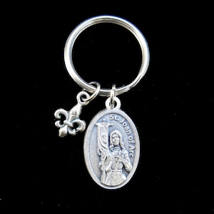May include: Silver keychain with a silver charm featuring a depiction of Saint Joan of Arc. The charm has the text "St. Joan of Arc" around the edge. The keychain also has a silver fleur-de-lis charm.