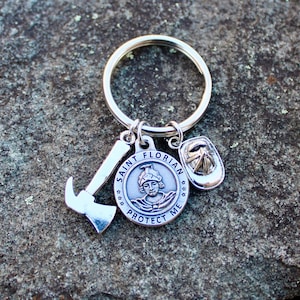 May include: Silver keychain with a Saint Florian medal, a firefighter's helmet charm, and an axe charm. The medal features a depiction of Saint Florian and the words "Saint Florian Protect Me".
