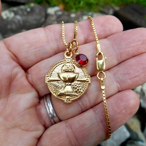 May include: Gold chain necklace with a gold chalice pendant and a red gemstone charm. The pendant features the word "JESUS" on the chalice.