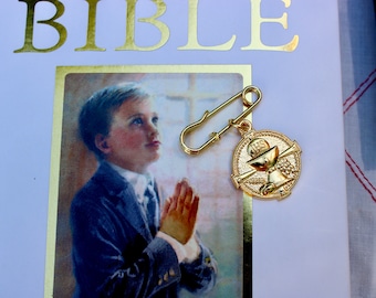 May include: A gold-colored pin with a religious symbol and a safety pin attached. The pin is on top of a book with the word "BIBLE" printed in gold letters.