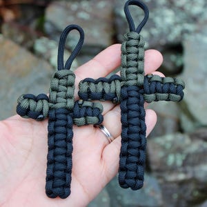 Puede incluir: Dos cruces artesanales de paracord negro y verde oliva. Cada cruz tiene una parte superior en bucle para colgar. Las cruces están hechas con un tejido apretado, mostrando la artesanía detallada. El fondo está borroso, enfatizando las cruces.