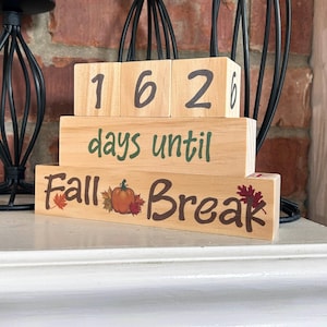 Könnte beinhalten: Holzblock-Kalender mit der Zahl "162" oben. Der mittlere Block zeigt "Tage bis" in Grün. Der untere Block zeigt "Herbstferien" mit einem Kürbis und Blättern.