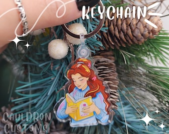 May include: A keychain featuring a cartoon illustration of a woman with red hair reading a book. The woman is wearing a blue sweater and the book is open to a page with a colorful illustration. The keychain is attached to a silver ring.