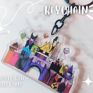 May include: A clear acrylic keychain featuring a colorful castle design with cartoon characters and Mickey Mouse balloons. The keychain has a black ring and the word "KEYCHAIN" is written in white. The text "CAULDRON CUSTOMS" is also visible.