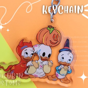 May include: A Halloween-themed keychain featuring three cartoon ducks. One duck is wearing a red devil costume, another is a ghost, and the third is wearing a blue witch hat. The keychain also includes a small orange pumpkin.