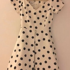 Puede incluir: Un vestido blanco con lunares negros y un escote en V. El vestido tiene mangas cortas y un corpiño ajustado. El vestido está hecho de una tela ligera y tiene una cremallera en la espalda.