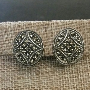 May include: A pair of silver earrings with a geometric design set with black marcasite stones. The earrings are displayed on a brown burlap background.