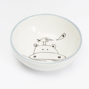 Puede incluir: Un tazón de cerámica blanco con un borde azul y una ilustración en blanco y negro de un hipopótamo con un pájaro en su espalda. El pájaro sostiene un corazón en su pico.