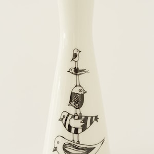 May include: A white ceramic vase with a flared shape, adorned with a whimsical illustration of stacked birds in black ink. The birds vary in design, with striped and patterned details. The vase is a decorative item.