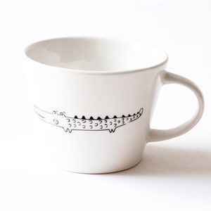 Pode incluir: Caneca de cerâmica branca com um desenho de linha preta de um crocodilo no lado.