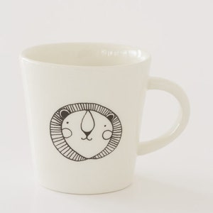 Könnte beinhalten: Cremefarbene Keramiktasse mit einer schwarz-weißen Löwengesicht-Illustration. Die Tasse hat einen gebogenen Henkel und ein schlichtes Design. Die Löwenillustration ist auf der Vorderseite der Tasse zentriert.
