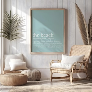 May include: A framed print with the words "the beach" in white text on a light blue background. The definition of "the beach" is also included. The print hangs above a wicker chair and a woven ottoman, creating a coastal theme.