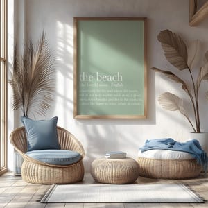 May include: A serene interior scene featuring a framed print with the words "the beach" and its definition. A wicker chair with a blue cushion and pillow, a round ottoman, and a woven footstool with a blue blanket complete the coastal decor.