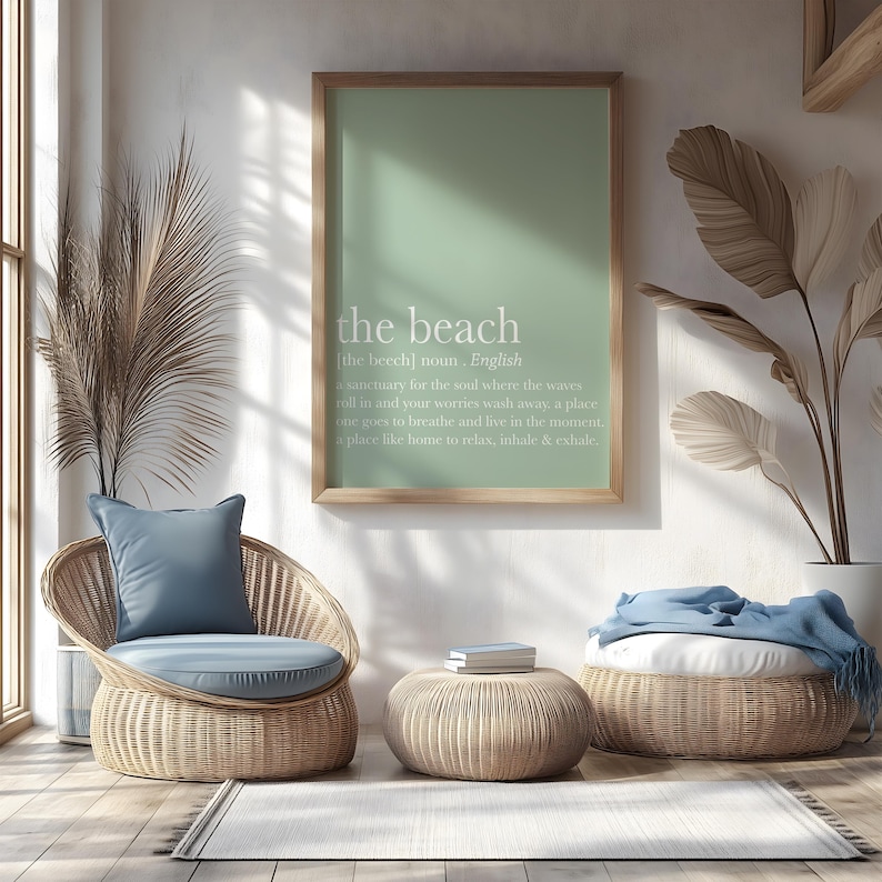 May include: A serene interior scene featuring a framed print with the words "the beach" and its definition. A wicker chair with a blue cushion and pillow, a round ottoman, and a woven footstool with a blue blanket complete the coastal decor.