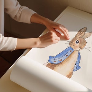 May include: A watercolour painting of Peter Rabbit wearing a blue jacket. The painting is on a white sheet of paper and is being rolled up by a person's hand.