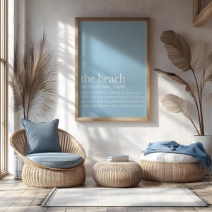 May include: A light blue framed print with the words "the beach" and a definition. The print hangs above a wicker chair with a blue cushion and pillow. A matching ottoman and a woven basket with a blue blanket are also in the room. The room has a beachy, coastal theme.