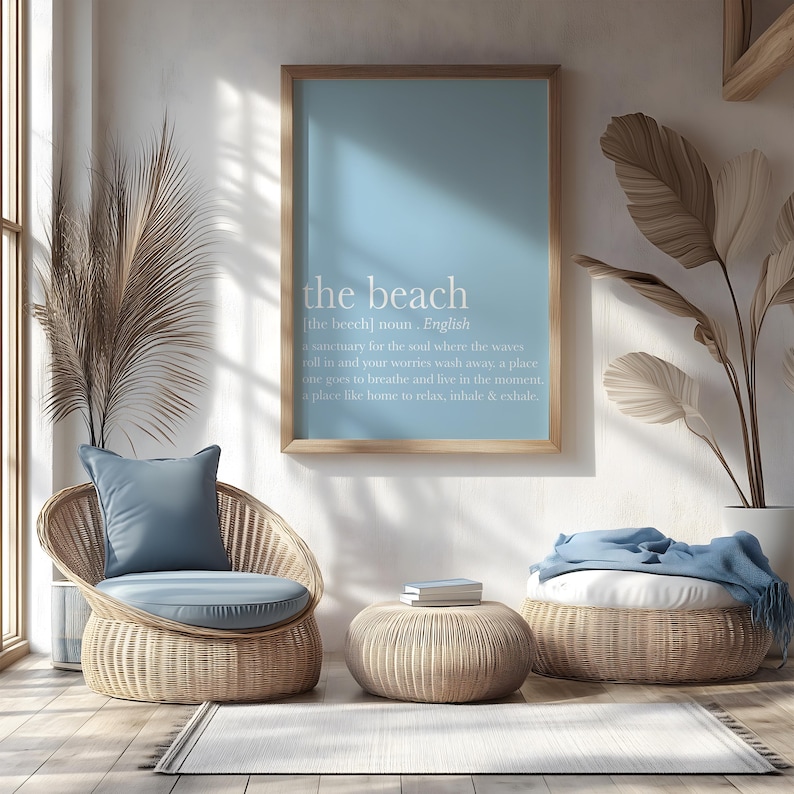 May include: A light blue framed print with the words "the beach" and a definition. The print hangs above a wicker chair with a blue cushion and pillow. A matching ottoman and a woven basket with a blue blanket are also in the room. The room has a beachy, coastal theme.