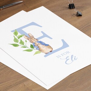May include: A blue letter E with a watercolour illustration of Peter Rabbit in a blue jacket. The letter is surrounded by green leaves. The text "E is for Eli" is written below the letter.