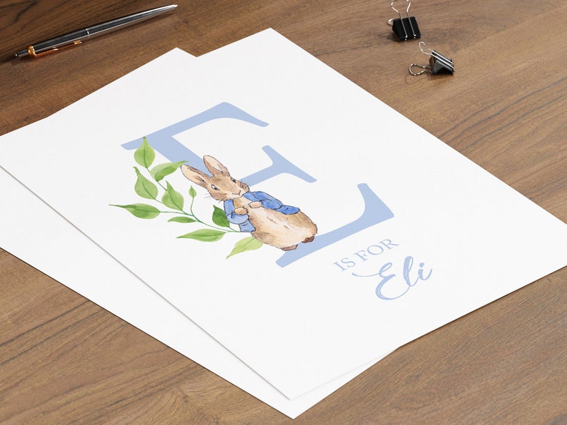 May include: A blue letter E with a watercolour illustration of Peter Rabbit in a blue jacket. The letter is surrounded by green leaves. The text "E is for Eli" is written below the letter.