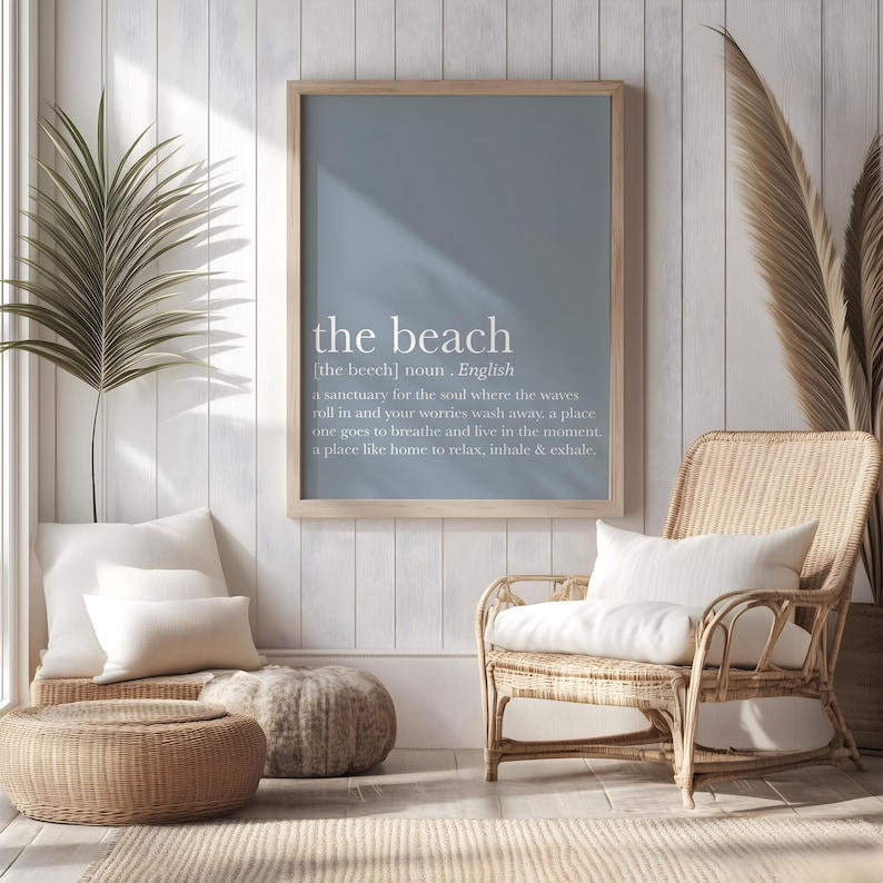 May include: A framed print with the text "the beach" in white, with a definition below. The print hangs above a wicker chair with a white cushion. A woven ottoman and pillows are in front of the chair. The room has a coastal theme.
