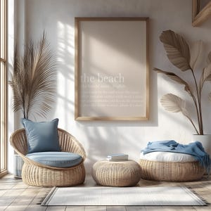 May include: A light-filled interior scene featuring a framed print with the text "the beach." Wicker furniture includes a chair with a blue cushion, a round ottoman, and a larger woven pouf. A rug and decorative plants complete the serene setting.