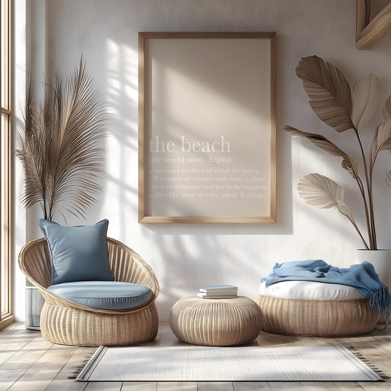 May include: A light-filled interior scene featuring a framed print with the text "the beach." Wicker furniture includes a chair with a blue cushion, a round ottoman, and a larger woven pouf. A rug and decorative plants complete the serene setting.
