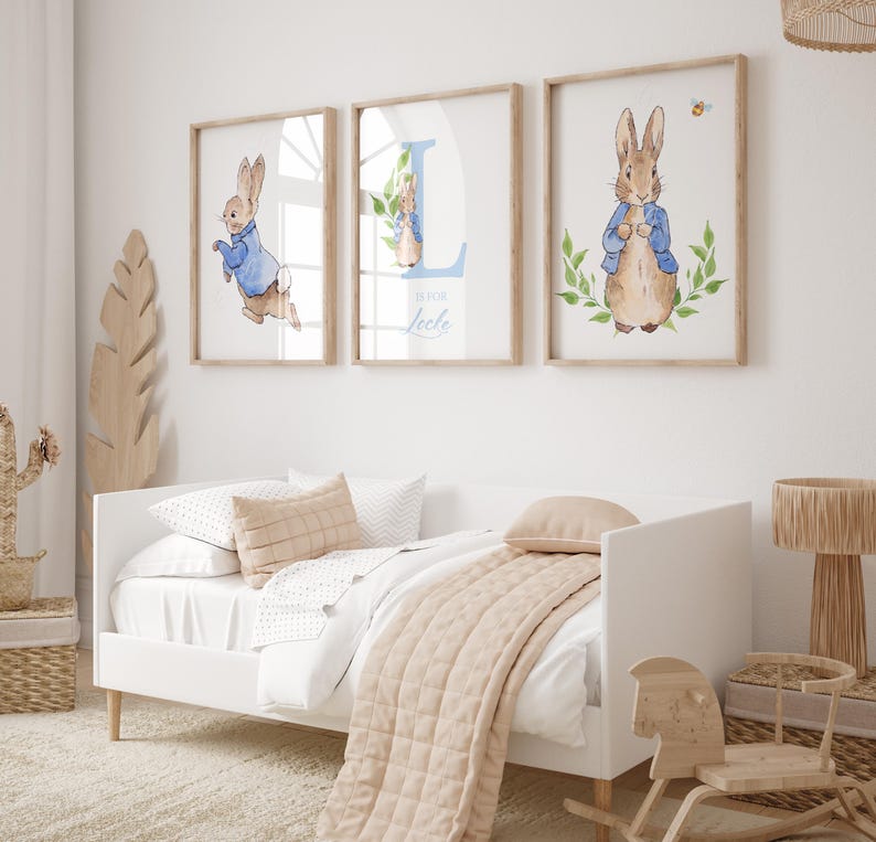 May include: Three framed prints featuring Peter Rabbit illustrations. The prints are in a nursery setting with a white daybed and a rocking horse. The prints feature a blue and white letter L, a Peter Rabbit in a blue jacket, and a Peter Rabbit in a blue jacket with a bee.