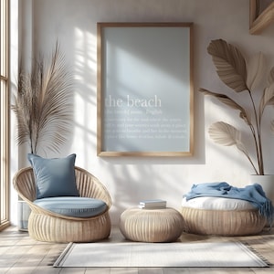 May include: A light-filled interior scene featuring a framed print with the text "the beach" above a wicker chair with a blue cushion and pillow. A small round table and a larger ottoman, both made of wicker, are also present. A rug and decorative plants complete the coastal theme.