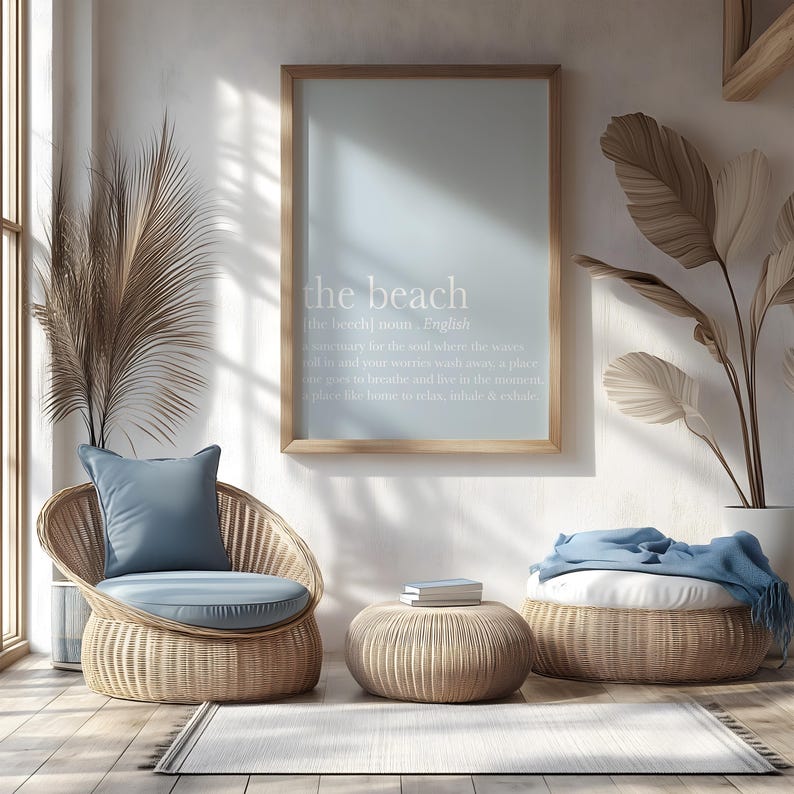 May include: A light-filled interior scene featuring a framed print with the text "the beach" above a wicker chair with a blue cushion and pillow. A small round table and a larger ottoman, both made of wicker, are also present. A rug and decorative plants complete the coastal theme.