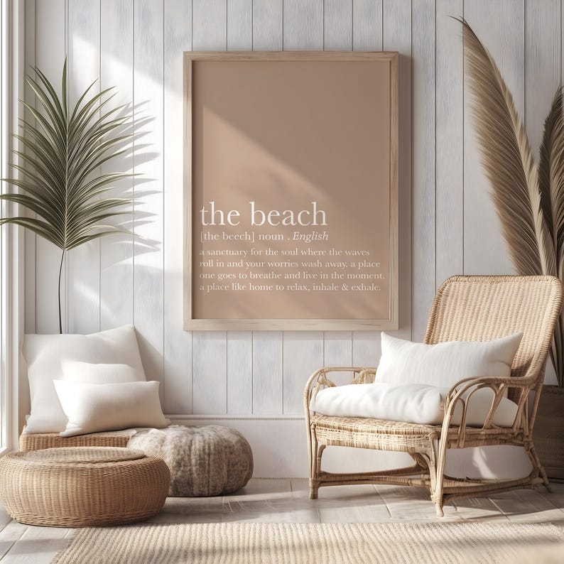 May include: A framed print with the text "the beach" in white, with a definition below. The print hangs above a wicker chair with a white pillow. Other decor includes a woven ottoman, pillows, and a palm frond.