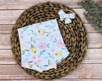 May include: A set of baby clothes featuring a floral print with pink and yellow flowers on a white background. The set includes a pair of bloomers and a matching headband.