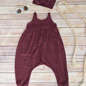 May include: A maroon sleeveless jumpsuit with a gathered waist and cuffed legs. The jumpsuit is laid flat on a woodgrain background with a headband, sunglasses, and a wooden bead necklace.