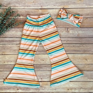 Puede incluir: Un pantalón de piernas anchas a rayas con una diadema a juego. El pantalón es de color rosa claro, marrón, turquesa y blanco a rayas. La diadema es de color rosa claro con un lazo marrón.