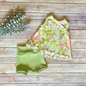 May include: A two-piece baby outfit featuring a floral print top and green shorts. The sleeveless top has a white ruffled hem and a pattern of pink, yellow, and green flowers. The shorts are a solid light green color.