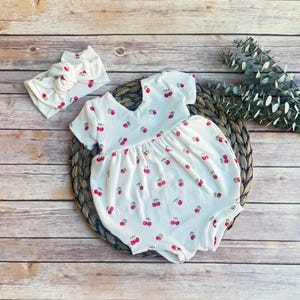 May include: White baby romper and matching headband, both adorned with a repeating cherry pattern. The romper features short sleeves and a gathered skirt, while the headband has a bow. The outfit is set on a woven mat.