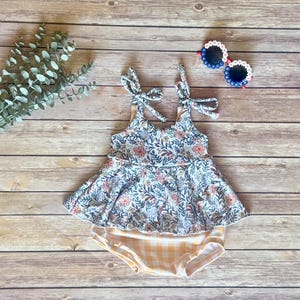 May include: A floral print baby swimsuit with tie straps. The top features a blue, pink, and green floral pattern, and the bottom is a yellow and white gingham print. A pair of sunglasses with white frames and blue lenses are also in the image.