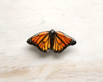 May include: A handcrafted monarch butterfly with orange and black wings, detailed with black outlines and white spots. The butterfly is centered on a white, textured surface, showcasing its intricate design.